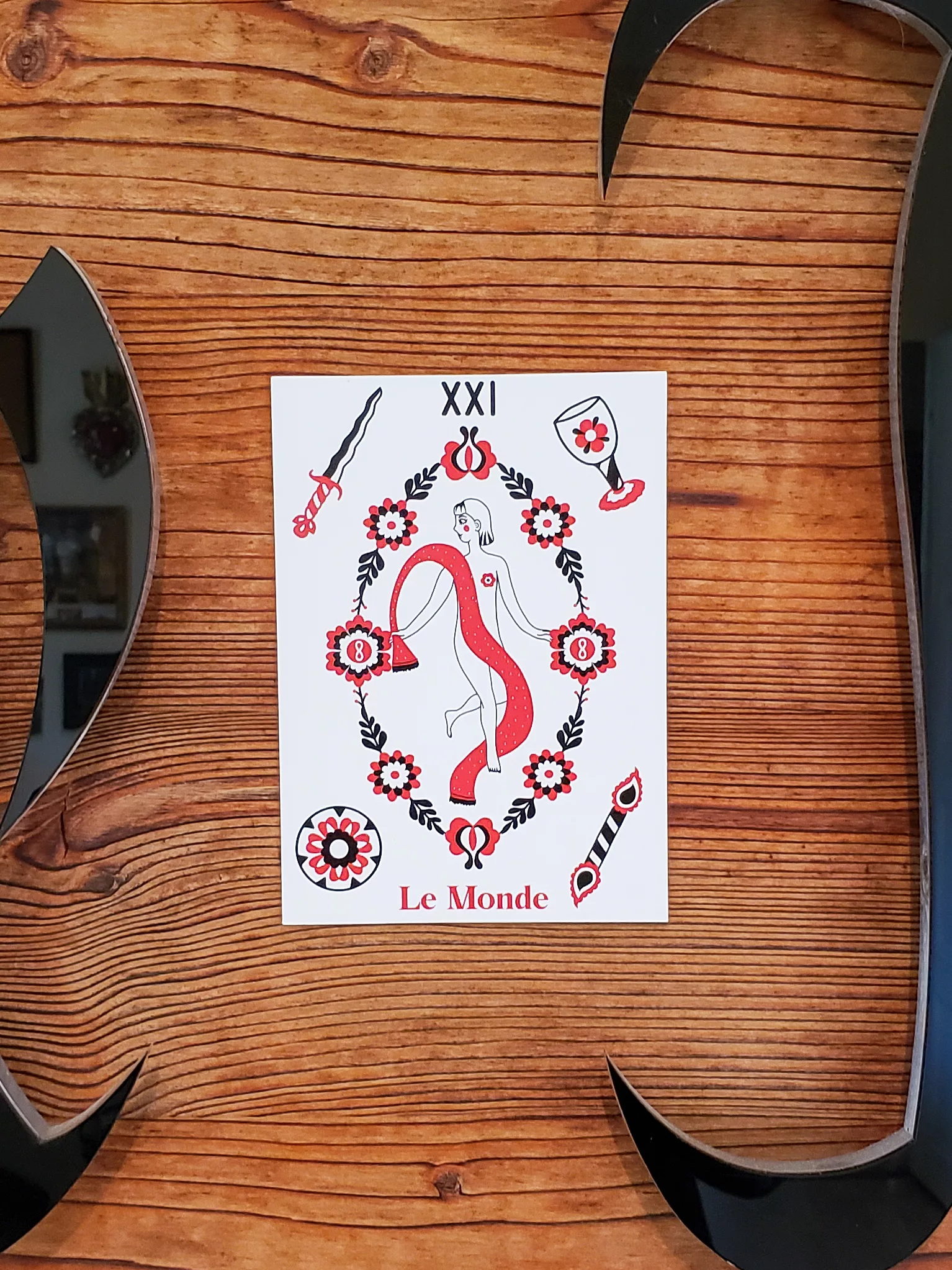 Carte postale illustrée de l'arcane Le Monde en rouge, noir et blanc avec motifs floraux, style pop folk Fortunae Studio